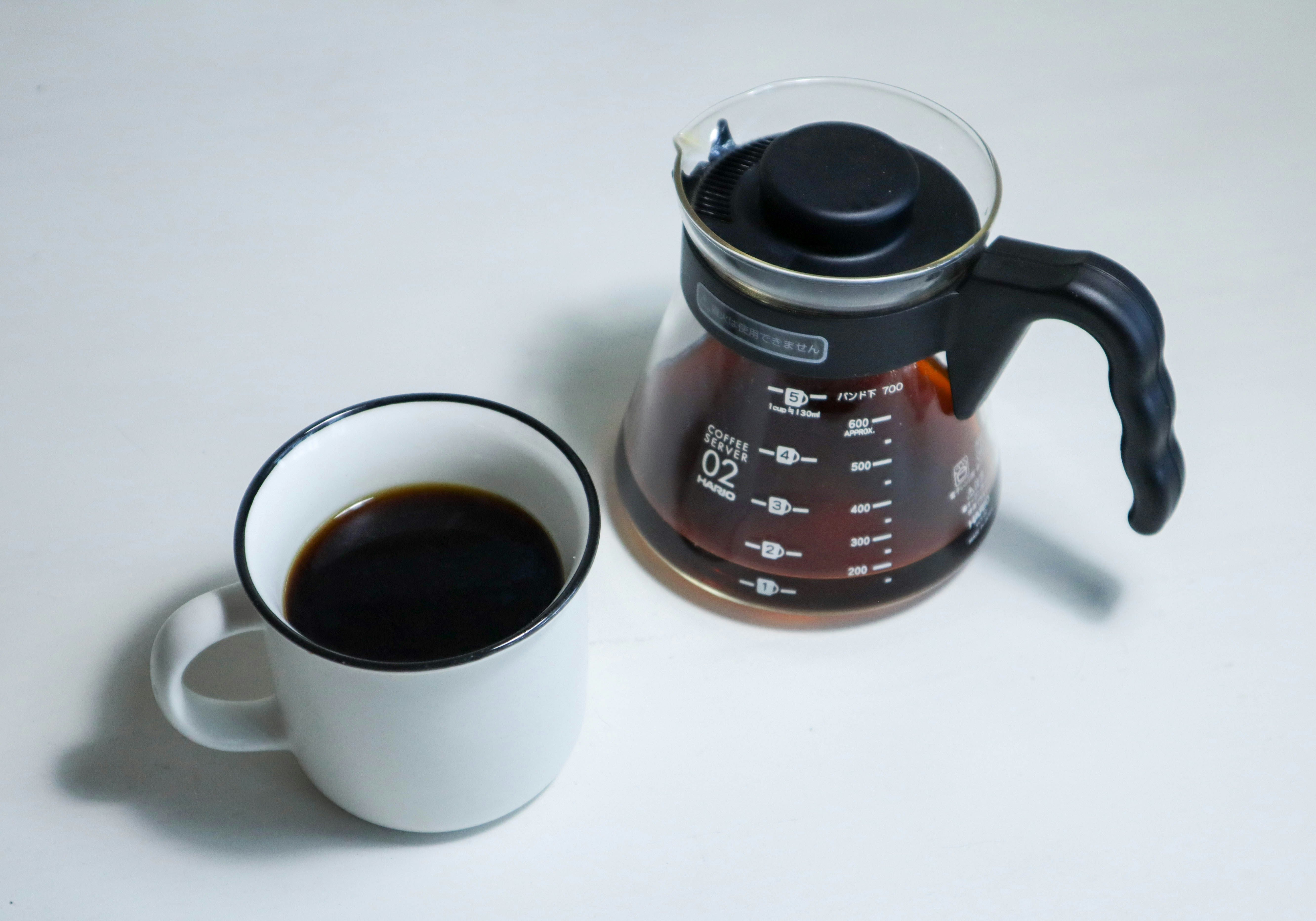 White mug filled with coffee next to a glass coffee carafe on a light gray background